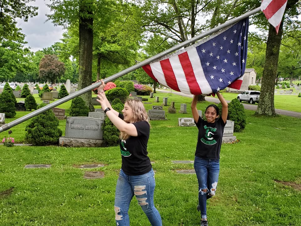 In a cemetery, two young women in matching black T-shirts and jeans smile as they lift an American flag on a metal pole.