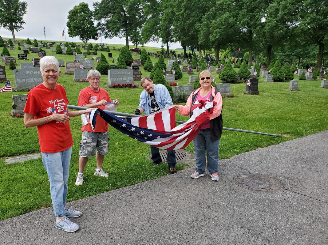 A group of four people folding an American flag, turned toward the camera and smiling, with gravestones in a cemetery in the background.