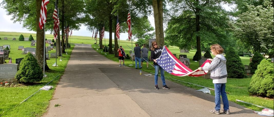 Standing on a paved lane in a cemetery, two women fold an American flags, while another group lowers a flag behind them.