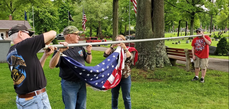 Two men and one woman in the foreground holding one end of a flag pole attaching a flag with another man in the background holding the other end of the pole on his shoulder.